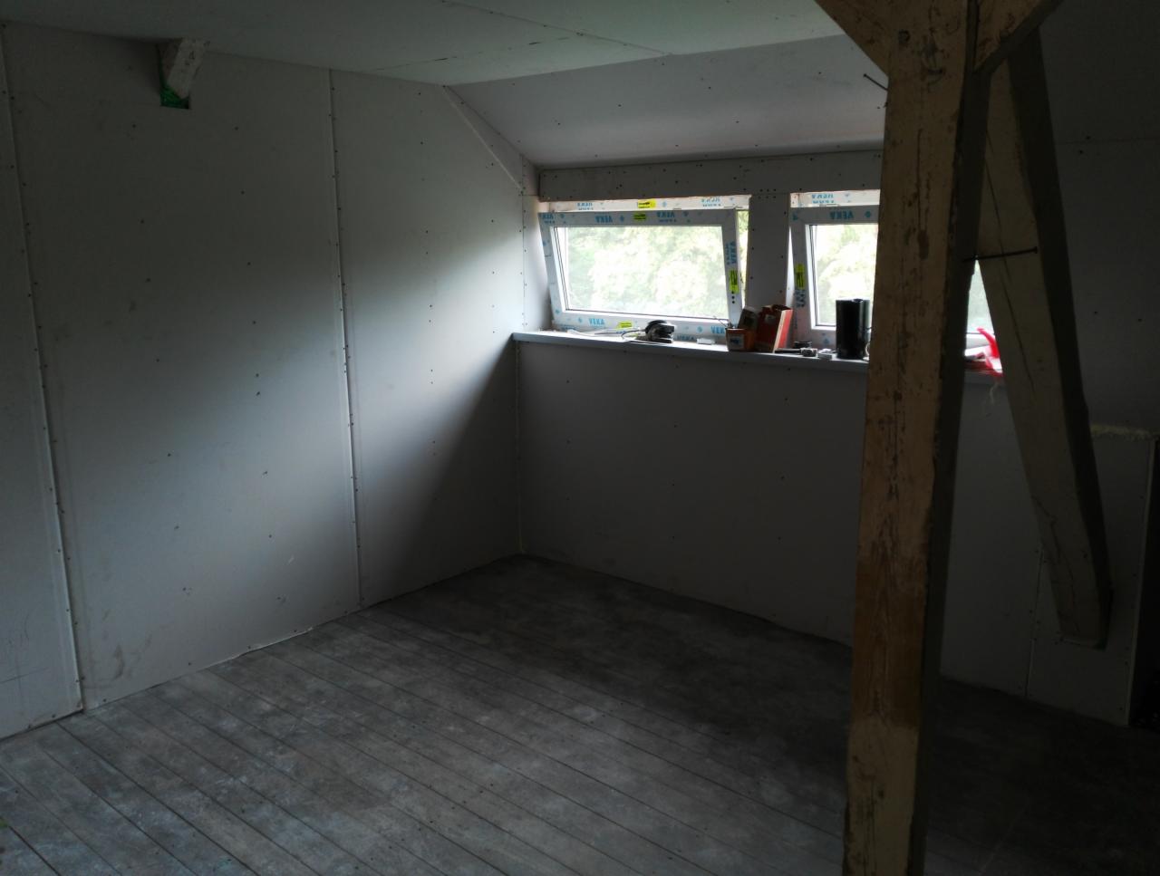 Pokój na poddaszu w trakcie prac wykończeniowych: ściany obłożone płytami gipsowo-kartonowymi, drewniana podłoga, okna dachowe z widokiem na zieleń, widoczna drewniana belka konstrukcyjna.