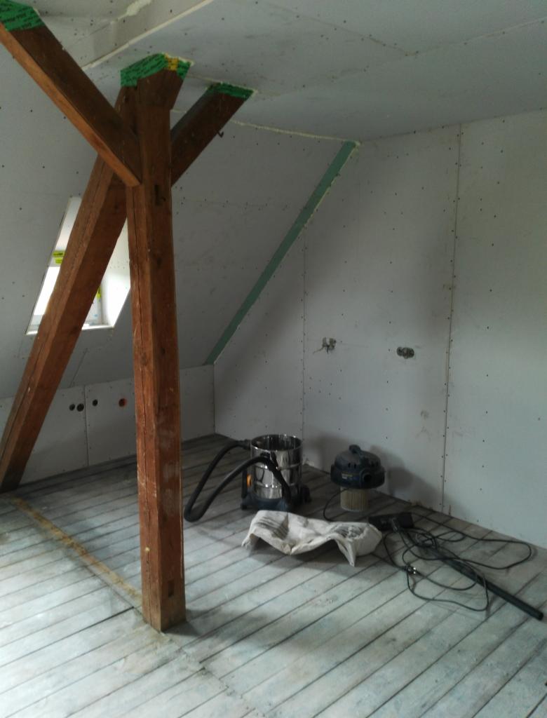 Pokój na poddaszu w trakcie remontu: widoczne drewniane belki, ściany wykończone płytami kartonowo-gipsowymi, szczeliny wypełnione pianką montażową, podłoga z desek, odkurzacz przemysłowy i biała...