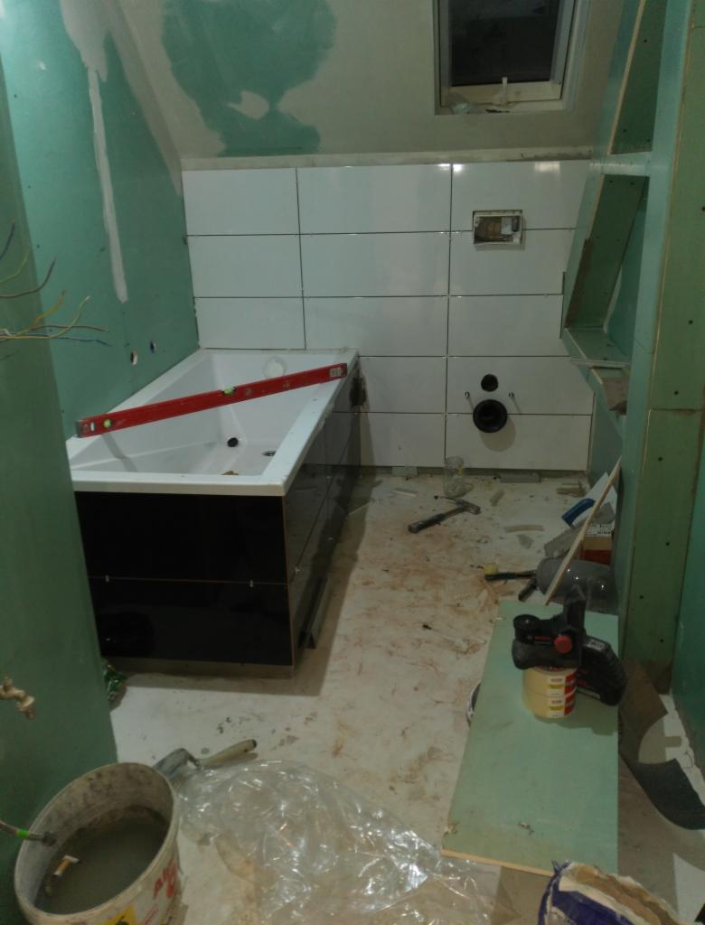 Łazienka w trakcie remontu: białe płytki na ścianach, czarna wanna z poziomicą, płyty karton gips na ścianach, narzędzia i materiały budowlane na podłodze.