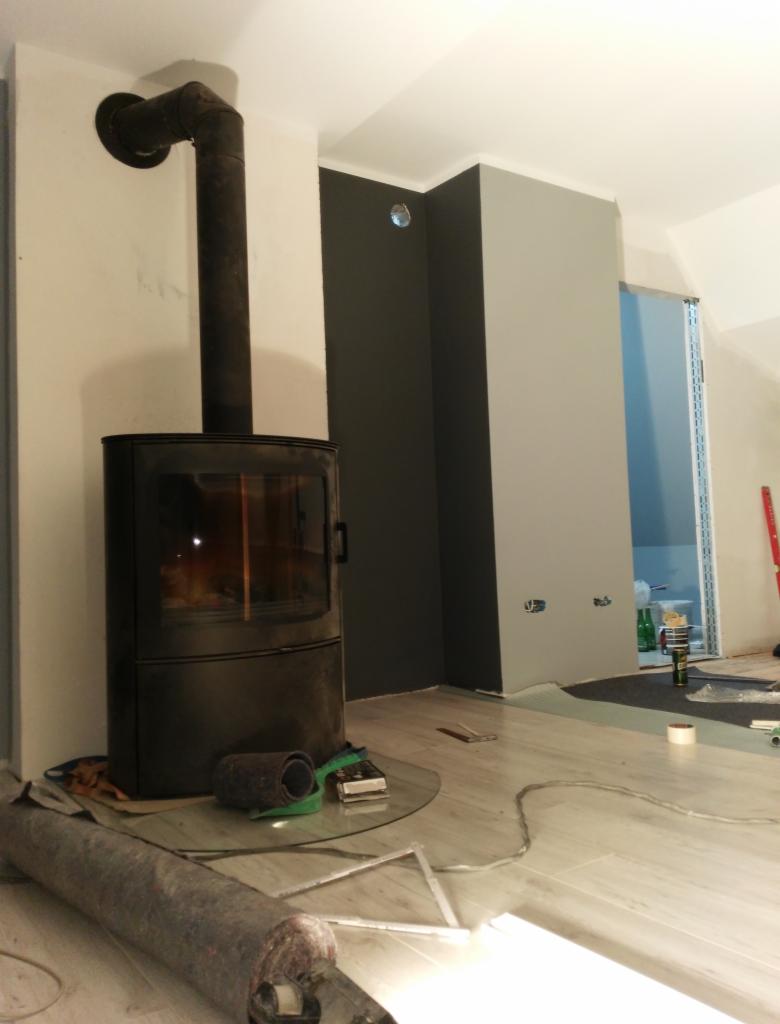 Czarny piec wolnostojący z podłączonym kominem w trakcie instalacji, widoczne narzędzia i materiały budowlane na podłodze, ściany pomalowane na różne odcienie szarości, częściowo zabezpieczone folią.