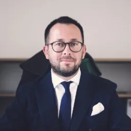 Portret adwokata w okularach, w ciemnym garniturze z poszetką i togą zawieszoną na krześle w tle.