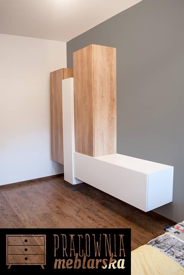 Nowoczesna szafa wisząca z drewnianymi elementami i białymi frontami, zamontowana na ścianie w pokoju z drewnianą podłogą, widoczne logo firmy meblarskiej w rogu.