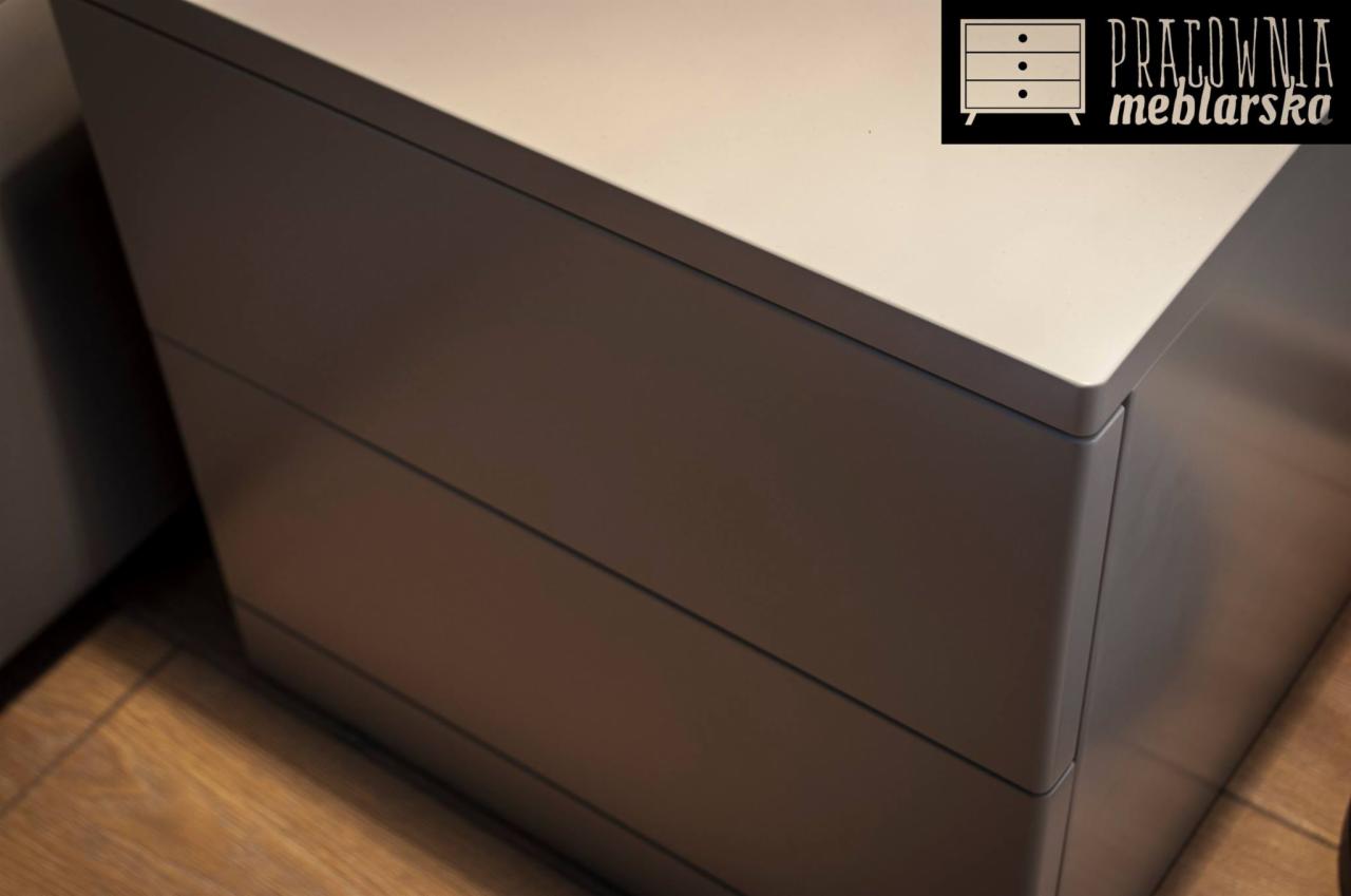 Szary stolik nocny z trzema szufladami, widoczny fragment podłogi i logo producenta w prawym górnym rogu.