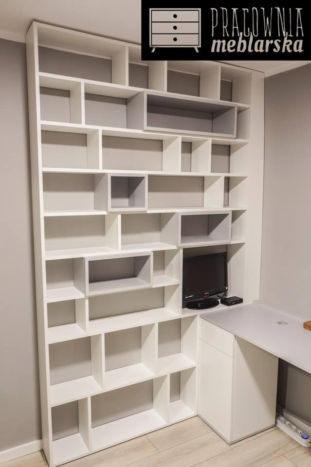 Biały regał z licznymi półkami o nieregularnym układzie, wbudowany w narożnik pokoju, z miejscem na telewizor i biurko obok.