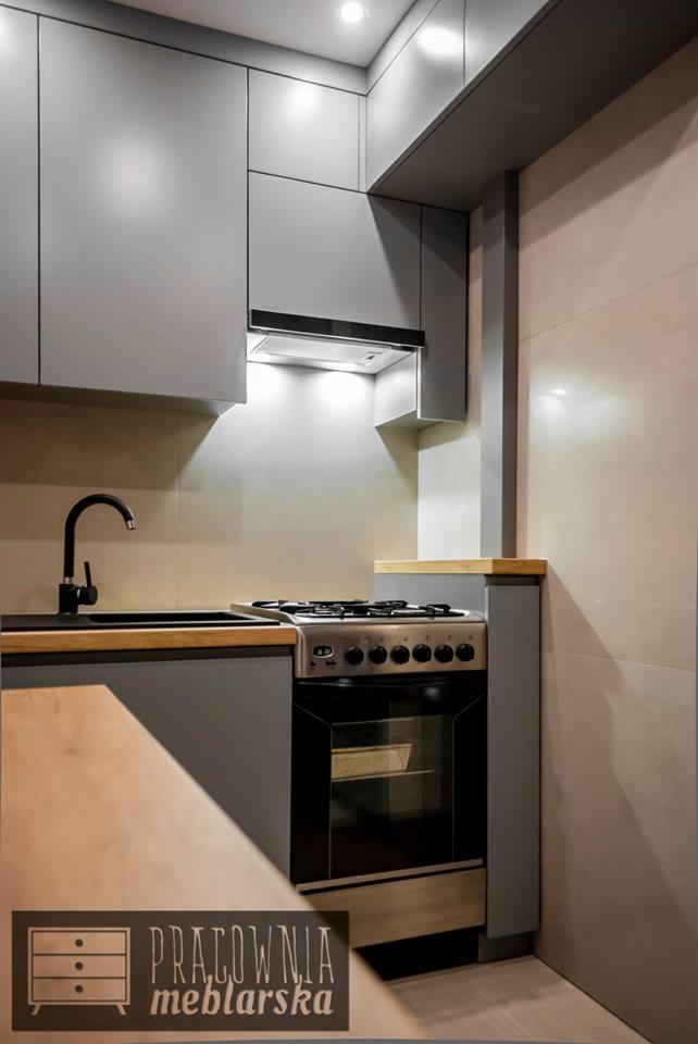 Narożnik kuchenny z szarymi szafkami, czarnym zlewem i kuchenką gazową; widoczny fragment blatu i logo pracowni meblarskiej w rogu.