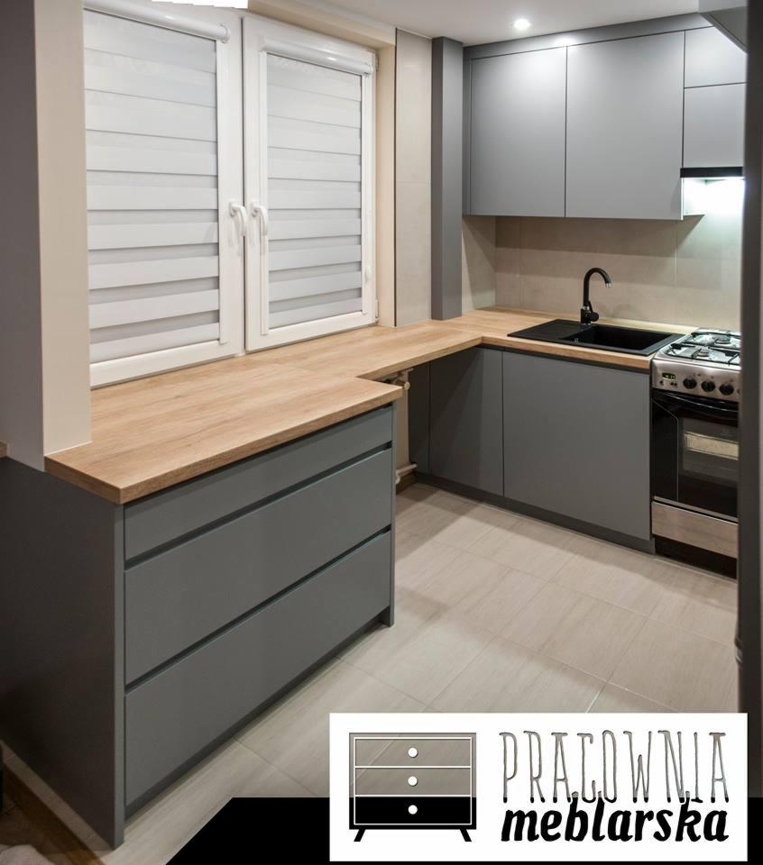 Szare meble kuchenne z drewnianym blatem pod oknem, czarny zlewozmywak i kuchenka gazowa, widoczne logo firmy meblarskiej w rogu.