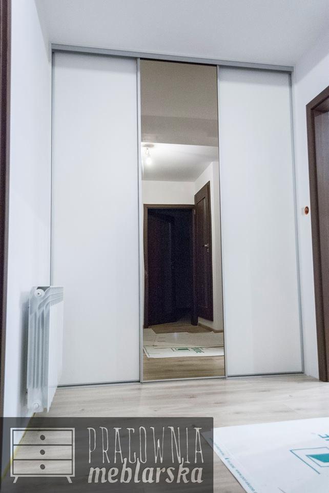 Biała szafa przesuwna z lustrem w przedpokoju, widoczne drzwi do pokoju i grzejnik, naklejka z logo firmy meblarskiej na dole zdjęcia.