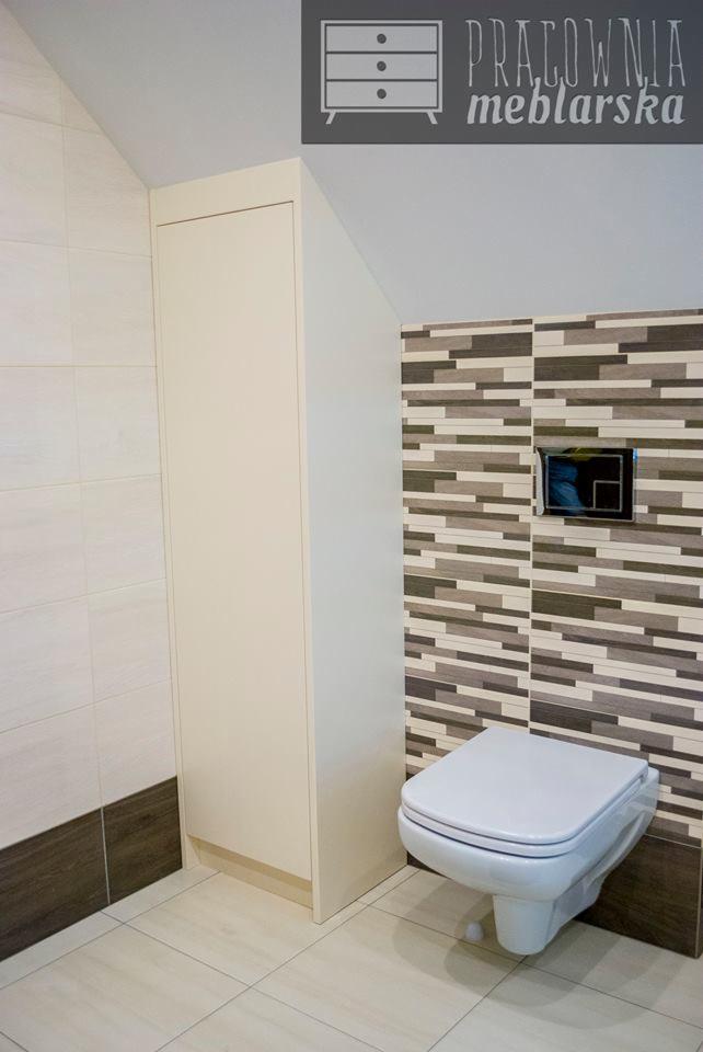 Wysoka, wąska szafka w zabudowie łazienki z płytkami w odcieniach beżu i brązu, wisząca toaleta w tle.