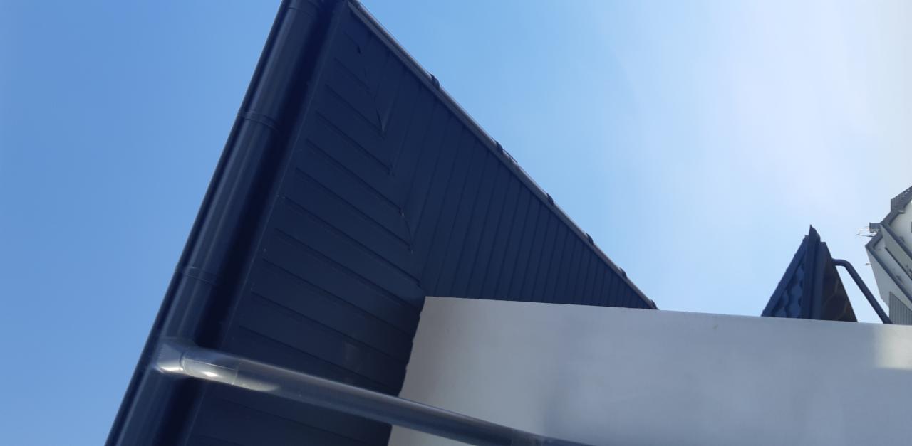 Fragment dachu z ciemną blachodachówką i orynnowaniem, widziany z dołu na tle jasnego nieba.