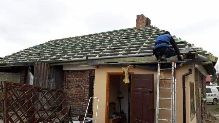 Pracownik w niebieskiej kamizelce na drabinie montuje folię dachową na drewnianej konstrukcji dachu jednopiętrowego budynku z ceglanym kominem.