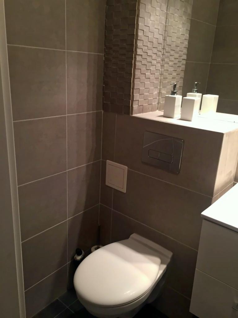 Nowoczesna toaleta w łazience z szarymi płytkami i lustrem z dekoracyjną ramą, biała armatura i dozowniki mydła.