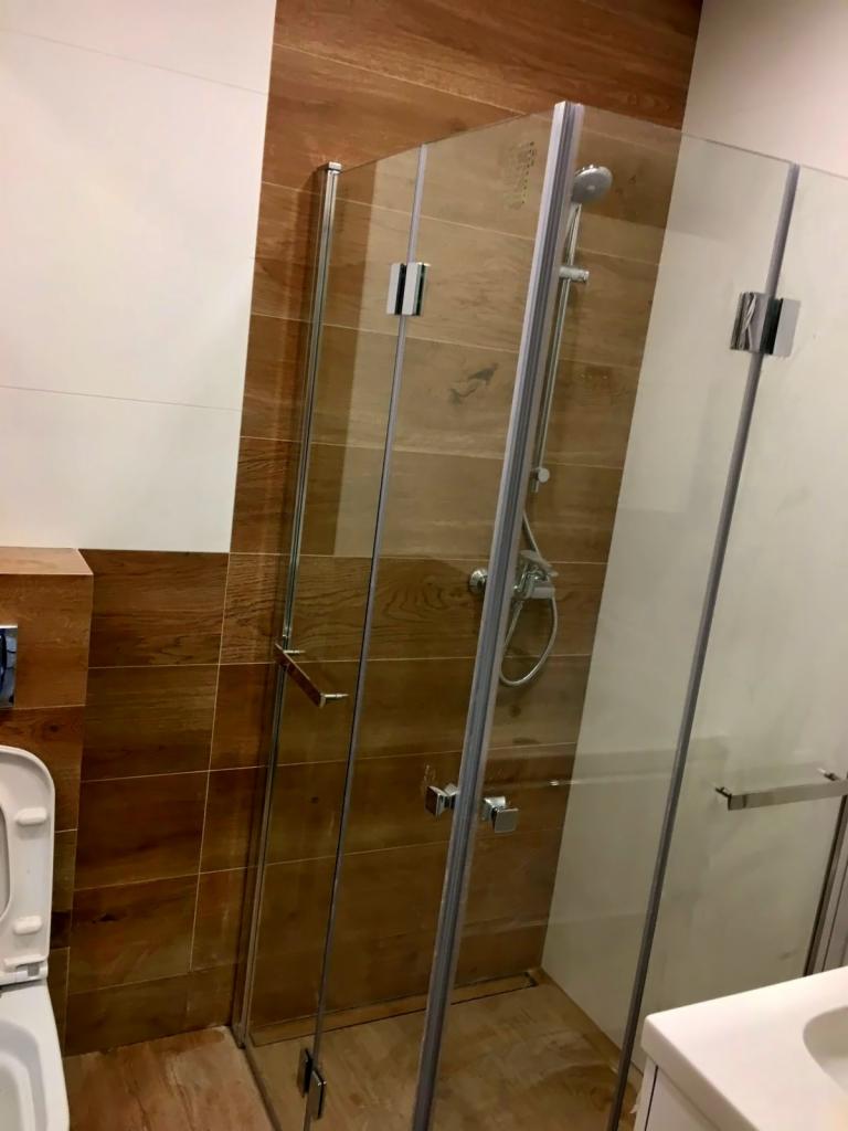 Nowoczesna kabina prysznicowa z transparentnego szkła, chromowanymi okuciami i deszczownicą, otoczona płytkami imitującymi drewno i białymi płytkami ściennymi, widoczna częściowo toaleta po lewej...