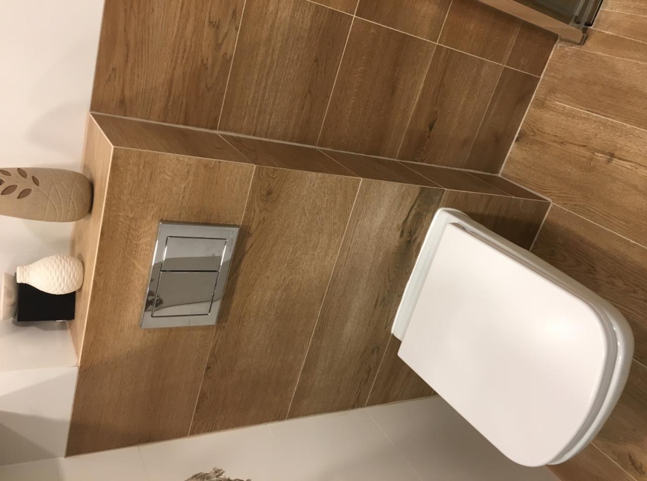 Nowoczesna łazienka z drewnopodobnymi płytkami, podwieszaną toaletą i chromowanym przyciskiem spłukującym.