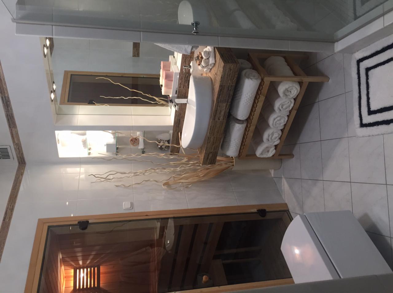 Nowoczesna łazienka z drewnianą umywalką, ręcznikami w rolkach na półce i widoczną sauną za szklanymi drzwiami.