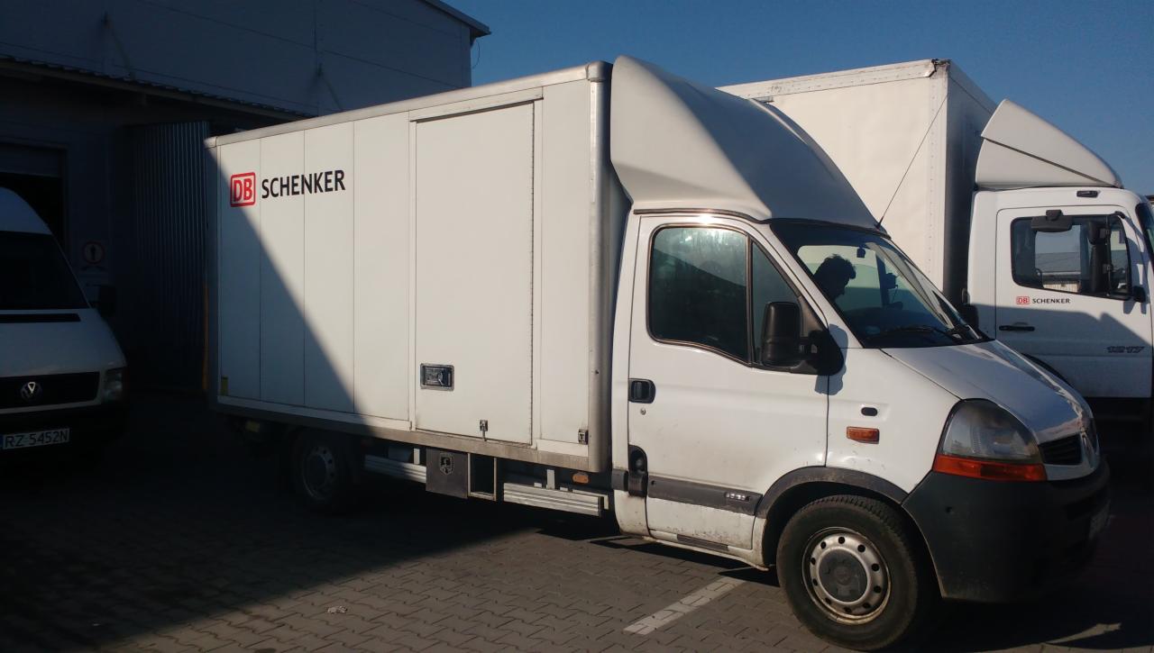 Biały samochód dostawczy z logo DB Schenker zaparkowany na placu, widoczny cień i fragment innego pojazdu w tle.