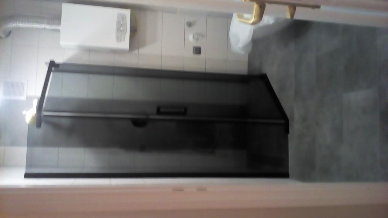 Szklana kabina prysznicowa z przesuwnymi drzwiami w łazience z szarymi płytkami, widoczny bojler na ścianie i fragment białej umywalki.