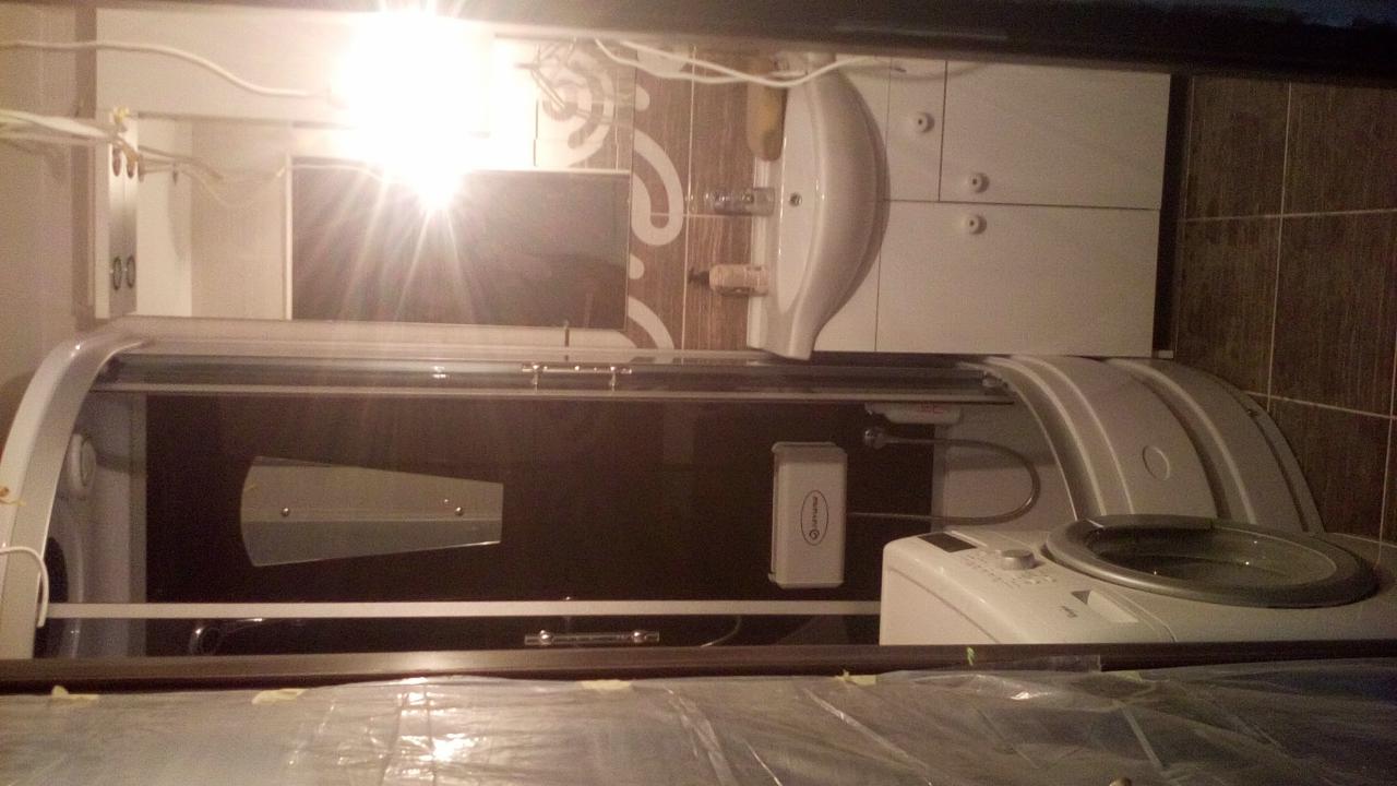 Nowoczesna kabina prysznicowa z hydromasażem w łazience z beżowymi płytkami, widoczna umywalka i pralka, całość oświetlona jasnym światłem.