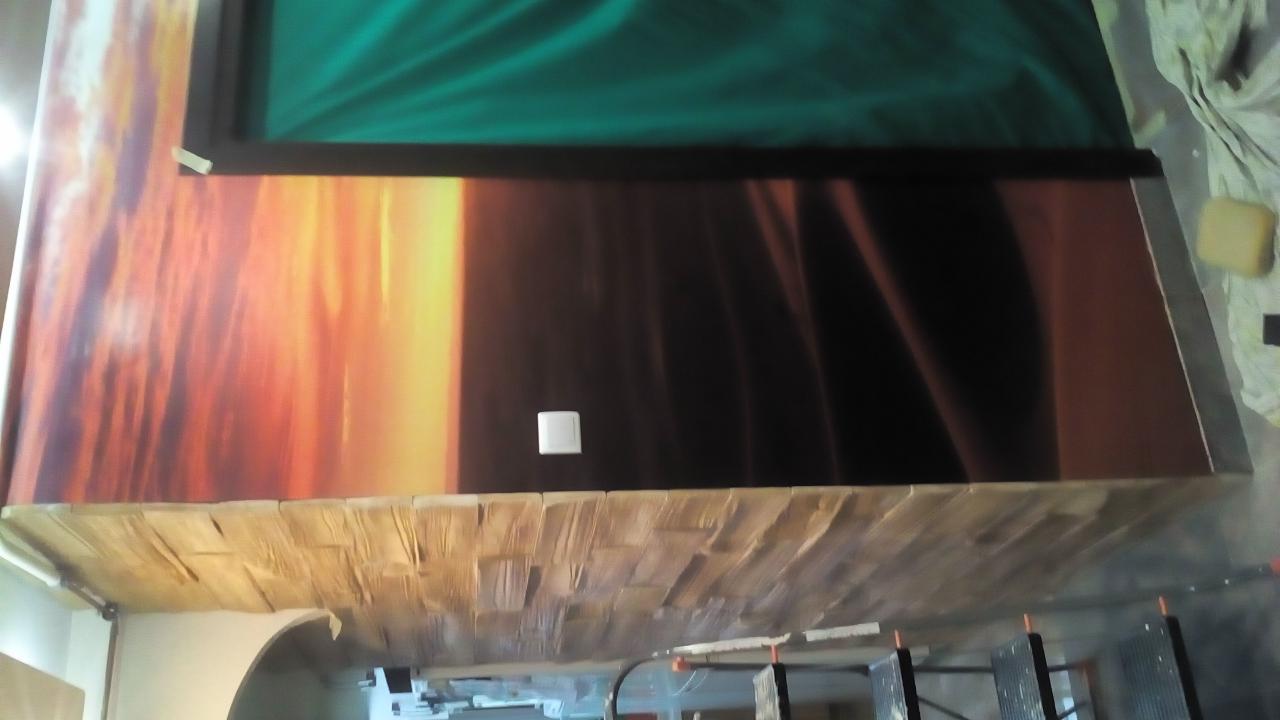 Ściana z fototapetą przedstawiającą zachód słońca nad morzem, wykończona drewnopodobną okładziną na dole, widoczny włącznik światła i fragment zielonego materiału u góry.