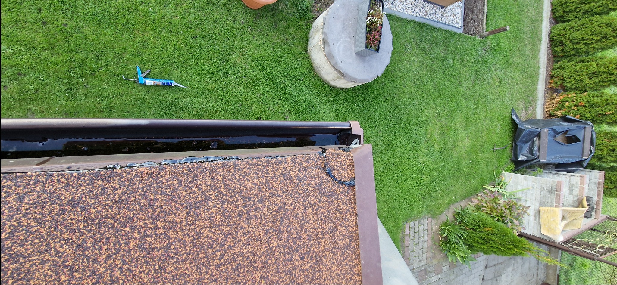 Uszczelnianie dachu z bliska: brązowy gont, rynna z wodą, narzędzie do silikonu na trawie. Widok z góry na naprawę dachu i otoczenie.