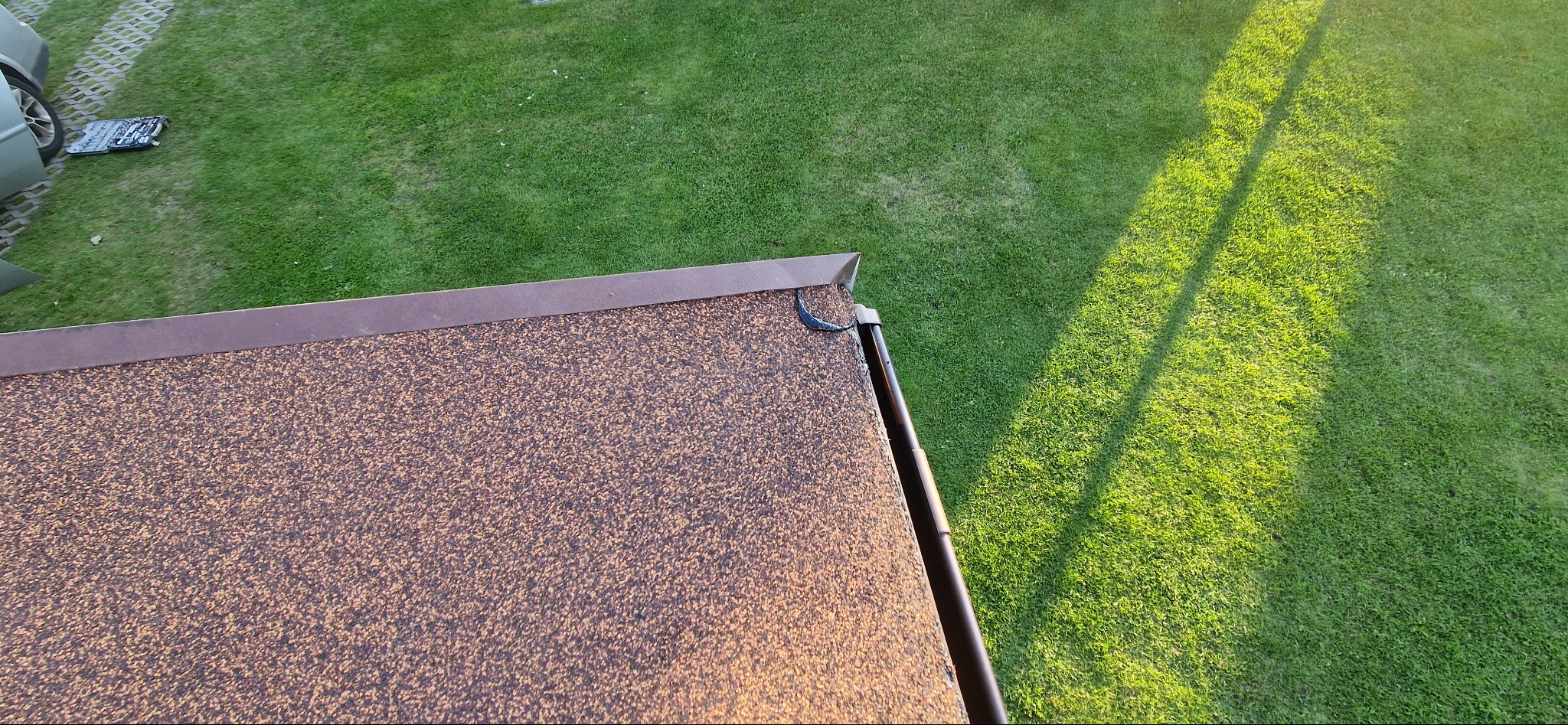 Widok z góry na brązowy dach pokryty gontem bitumicznym, z widocznym fragmentem rynny i trawnika w tle, oświetlonego popołudniowym słońcem.