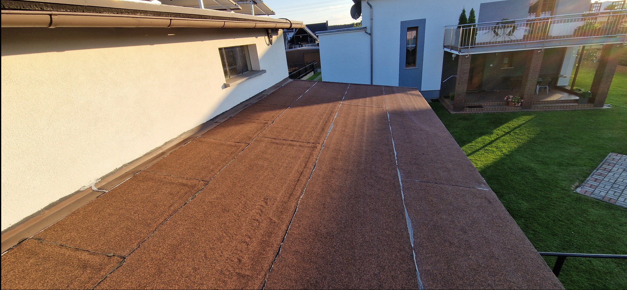 Dach pokryty brązową papą termozgrzewalną, widok z góry. Na drugim planie budynek z balkonem i zadbany trawnik w słoneczny dzień. Detal pokrycia dachowego.