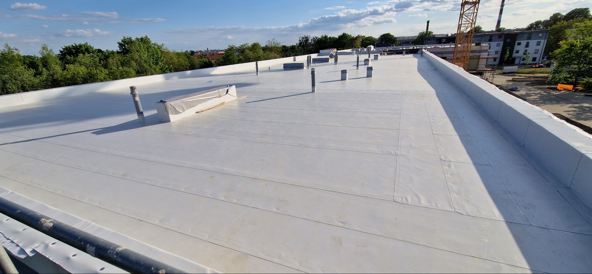 Biały, płaski dach budynku z widocznymi kominami wentylacyjnymi i obróbkami blacharskimi. W tle zieleń drzew i budynki mieszkalne, a w oddali żółty dźwig budowlany.