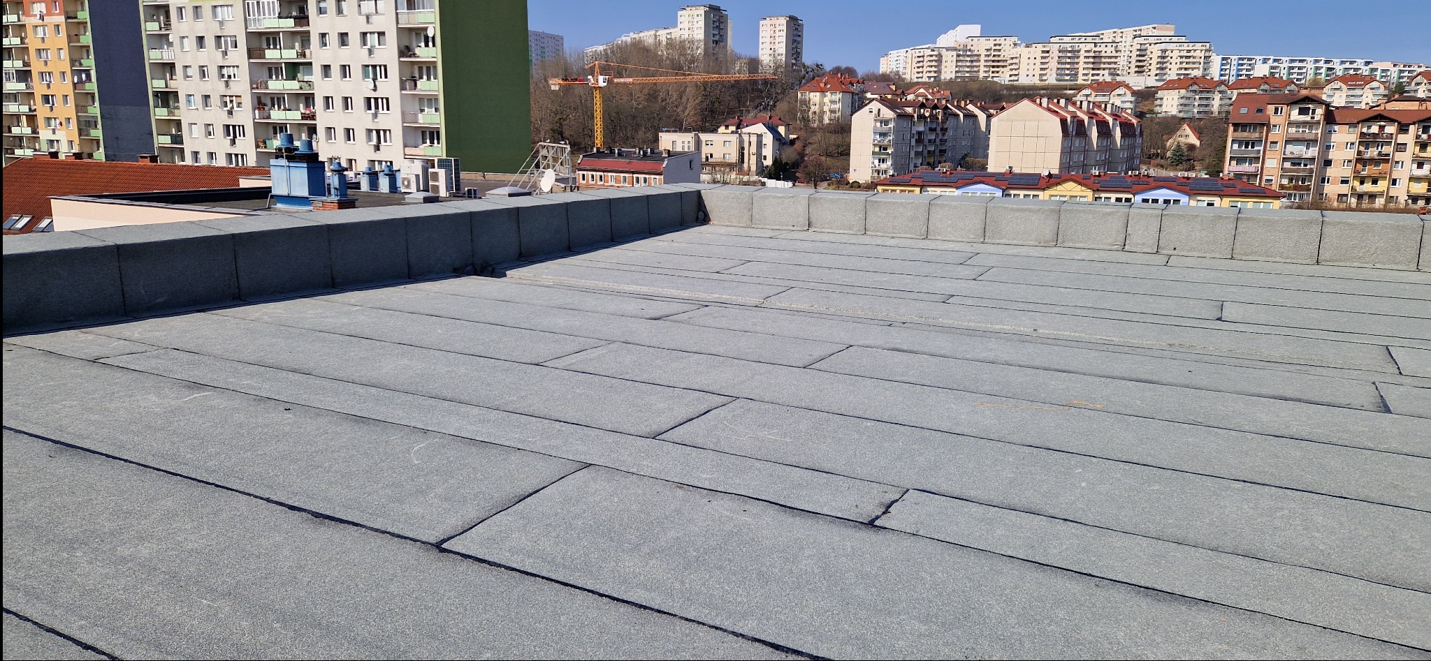 Szary, płaski dach pokryty papą na tle miejskiego krajobrazu z blokami i żurawiem budowlanym w oddali, w słoneczny dzień. Widok z góry.