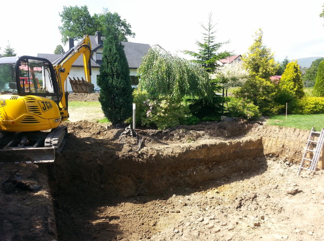 Żółta koparka JCB przy wykopie w ziemi, obok drzewa i krzewy, w tle dom jednorodzinny z ciemnym dachem i drabina oparta o skarpę wykopu.