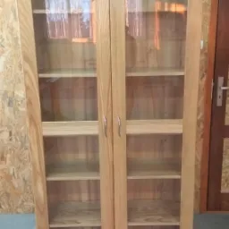 Dwudrzwiowa, przeszklona szafka z jasnego drewna z widocznymi półkami wewnątrz, ustawiona na szarym podłożu na tle ściany z boazerii i fragmentu ciemnych drzwi.