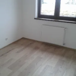 Jasne panele podłogowe ułożone w pokoju z białymi ścianami, grzejnikiem pod oknem z brązową ramą i widokiem na drzewa.