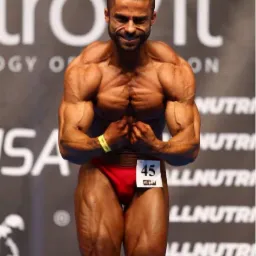 Kulturysta prezentuje umięśnienie na scenie konkursowej, z numerem 45, w czerwonych spodenkach, z widocznym logo sponsora w tle.