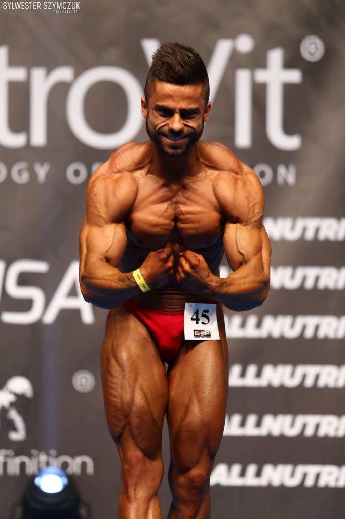 Kulturysta prezentuje umięśnienie na scenie konkursowej, z numerem 45, w czerwonych spodenkach, z widocznym logo sponsora w tle.
