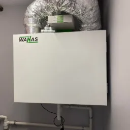 Biała centrala wentylacyjna z logo WANAS, podłączona do rur wentylacyjnych w pomieszczeniu technicznym.