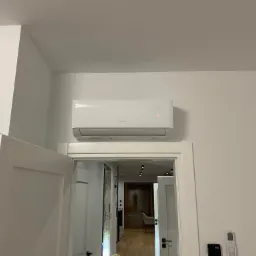 Biały klimatyzator marki Gree zamontowany nad futryną drzwi w jasnym pomieszczeniu, widoczny czujnik ruchu na ścianie i pilot klimatyzacji.