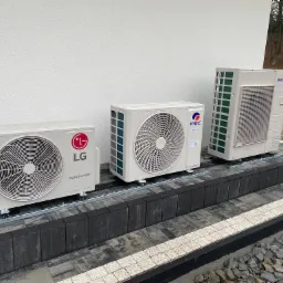 Trzy zewnętrzne jednostki klimatyzacyjne różnych producentów (LG, Gree, Samsung) ustawione na podwyższeniu z kostki brukowej przy ścianie budynku.