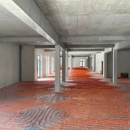 Surowy stan: Pomieszczenie w trakcie instalacji ogrzewania podłogowego z pomarańczowych rur na metalowej siatce, widoczne betonowe filary i ściany w stanie deweloperskim.