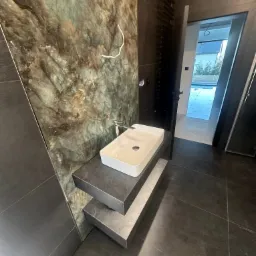 Nowoczesna łazienka z umywalką nablatową na dwupoziomowym blacie z płytek, ściana wyłożona płytkami imitującymi zielony marmur, widoczny grzejnik drabinkowy i otwarte drzwi do innego pomieszczenia.