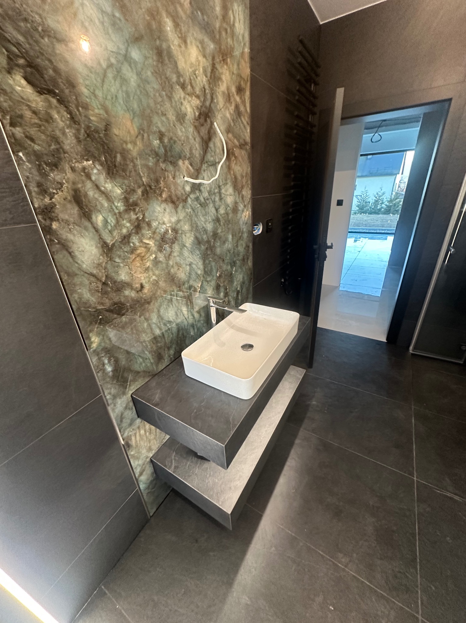 Nowoczesna łazienka z umywalką nablatową na dwupoziomowym blacie z płytek, ściana wyłożona płytkami imitującymi zielony marmur, widoczny grzejnik drabinkowy i otwarte drzwi do innego pomieszczenia.