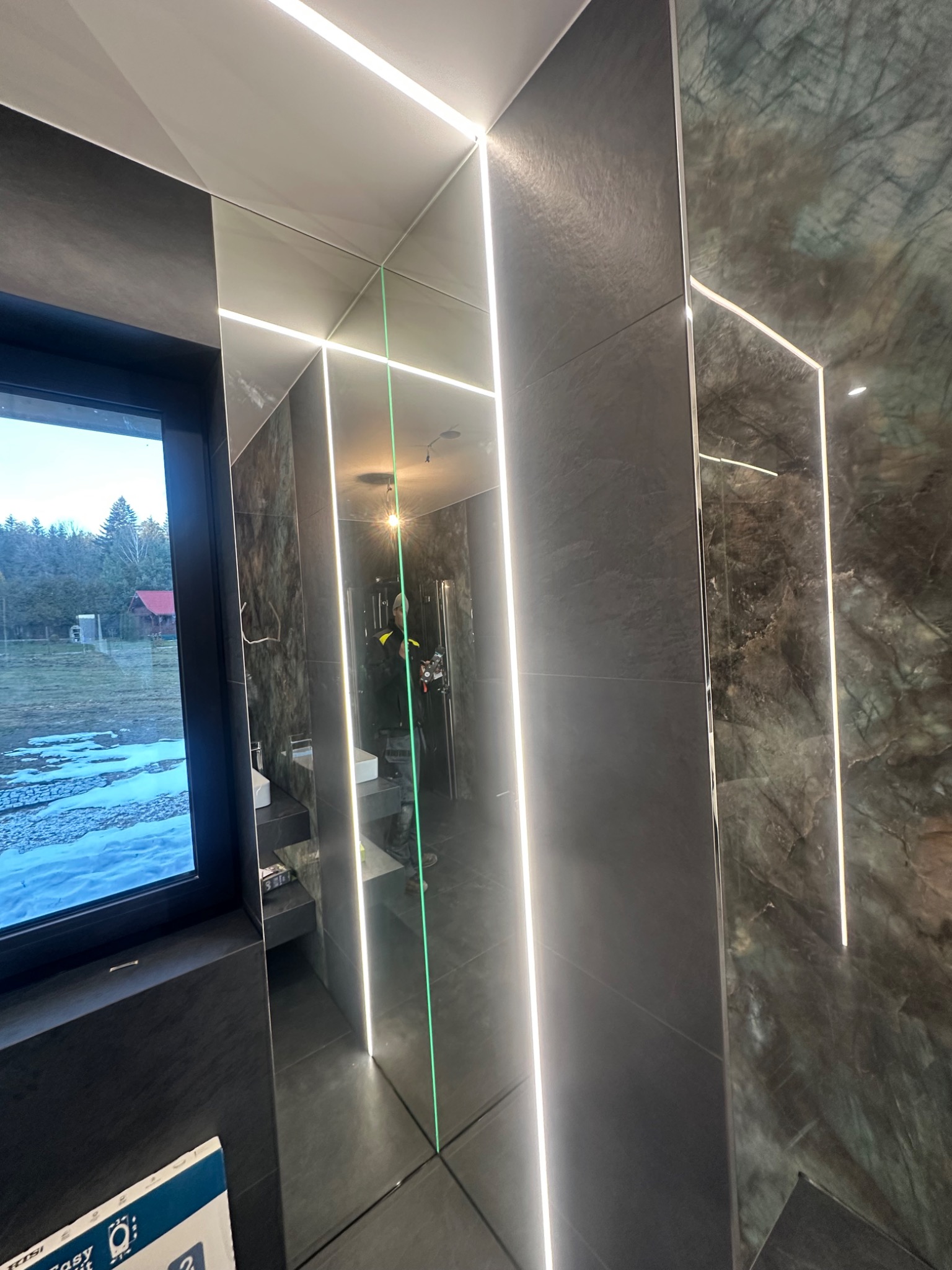 Nowoczesna łazienka z ciemnymi płytkami imitującymi kamień, podświetleniem LED wzdłuż krawędzi, dużym oknem z widokiem na zimowy krajobraz oraz odbiciem osoby pracującej wewnątrz.