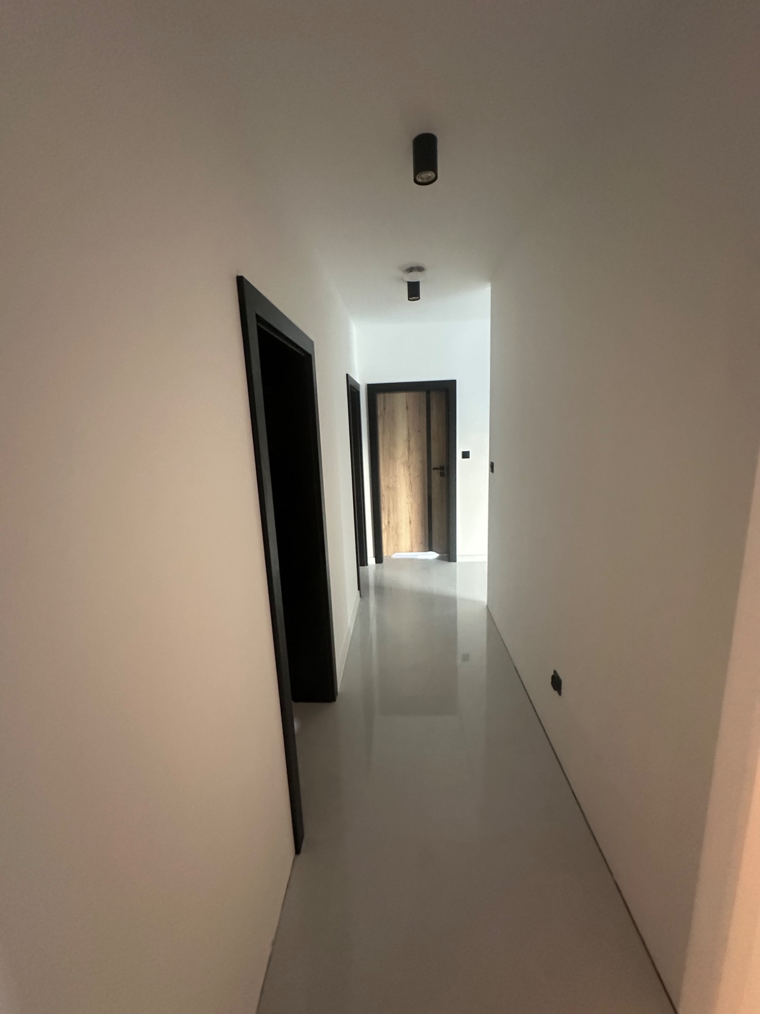 Wnętrze korytarza z szarą posadzką, białymi ścianami i czarnymi ościeżnicami drzwiowymi, widok w głąb mieszkania.