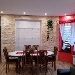 Jadalnia z dekoracyjną ścianą z kamienia imitującego piaskowiec, podświetlaną wnęką z szklanymi półkami, drewnianym stołem i krzesłami oraz czerwonym akcentem na ścianie z oknem.