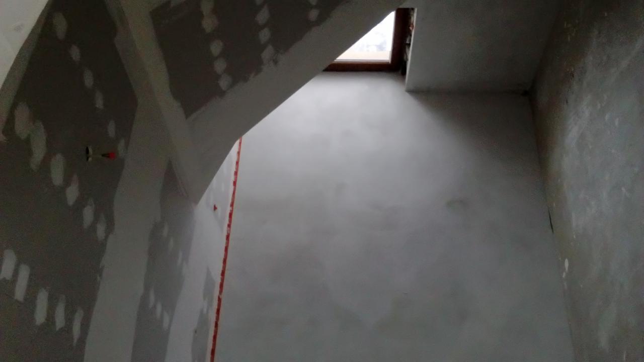 Poddasze w trakcie prac wykończeniowych: ściana ze skośnym sufitem częściowo pokryta płytami kartonowo-gipsowymi, na których widoczne są miejsca szpachlowania, okno dachowe w górnym rogu.