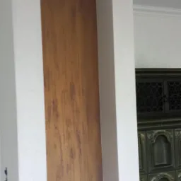 Dekoracyjna ściana z efektem drewna w niszy z białymi kolumnami i ozdobnym gzymsem, obok widoczny fragment pieca kaflowego.