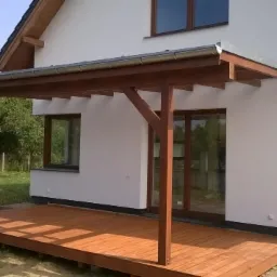 Zadaszony drewniany taras przylegający do białego domu z dużymi oknami, widoczne rynny i elementy konstrukcyjne dachu.