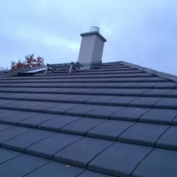 Dach pokryty ciemnoszarą dachówką ceramiczną, widoczny komin i fragment okna dachowego z zabezpieczeniem przeciwśniegowym, pochmurne niebo.