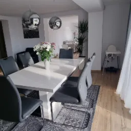 Jasna jadalnia z białym stołem, szarymi krzesłami, kwiatami w wazonie i srebrnymi lampami wiszącymi, widok na salon w tle, drewniana podłoga i dywan z wzorem.