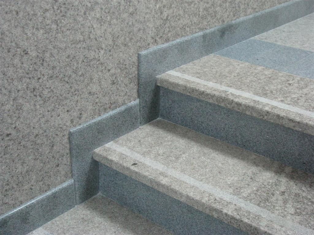 Granitowe schody z szarymi stopnicami i podstopnicami, z delikatnym białym paskiem antypoślizgowym na krawędziach, dopasowane kolorystycznie do ściany wykończonej tym samym materiałem.