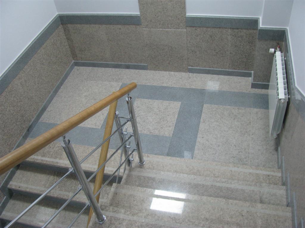 Kamienne schody z metalową balustradą i drewnianym poręczą, widok z góry, jasne płytki podłogowe z ciemniejszymi wstawkami, grzejnik na ścianie.