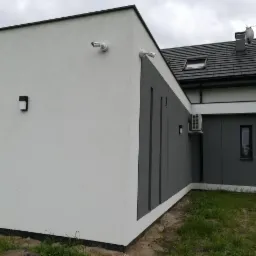 Nowoczesny dom z elewacją w odcieniach szarości i bieli, wyposażony w dwie kamery monitoringu, klimatyzator, oświetlenie zewnętrzne i antenę satelitarną na dachu.