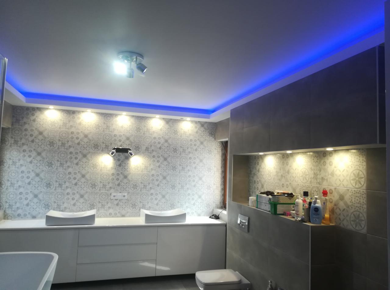 Nowoczesna łazienka z podświetleniem LED na suficie, wzorzystymi płytkami na ścianie, dwiema umywalkami nablatowymi i wnęką z oświetleniem punktowym oraz kosmetykami.